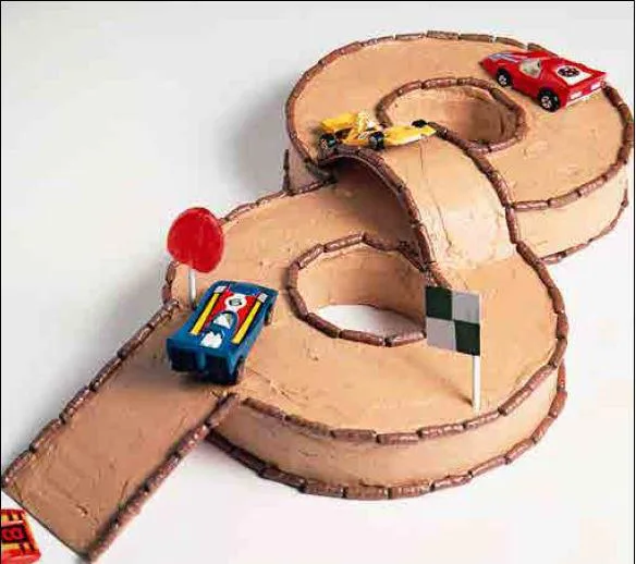 Race track-shaped cake with toy cars, lollipop and flag decorations, resembling a figure-eight track.