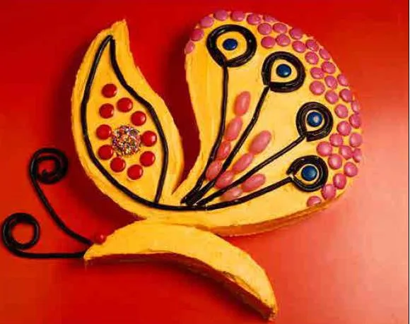 Butterfly-shaped cake with vibrant yellow frosting, black licorice outlines, and colorful candies on a red background.