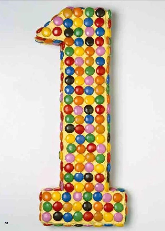 Candy-covered cake shaped like the number one, decorated with colorful round candies on a light background.