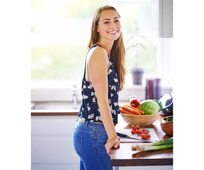 Smiling woman in kitchen with fresh vegetables on counter, wearing a floral top and jeans.