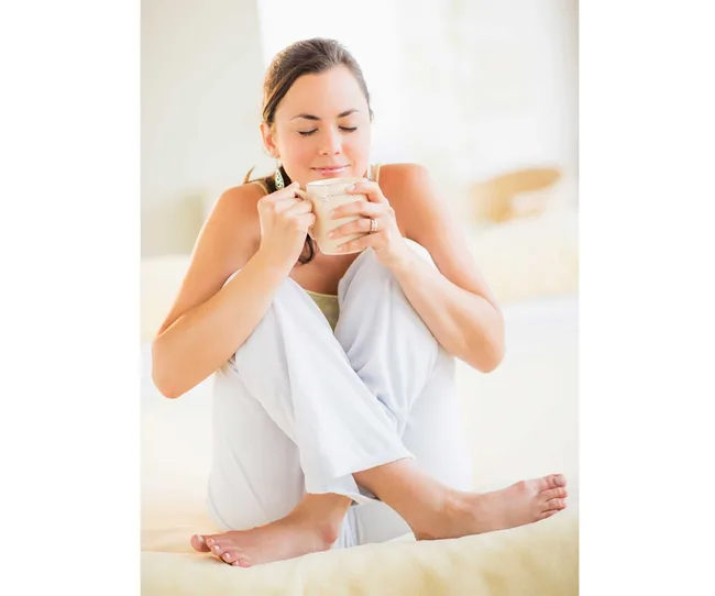 Woman sitting cross-legged, holding a cup with eyes closed and smiling, dressed in white, indoors.