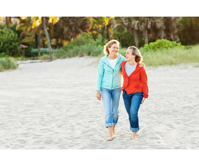 Two women smiling and walking barefoot on a sandy beach, surrounded by greenery and palm trees.