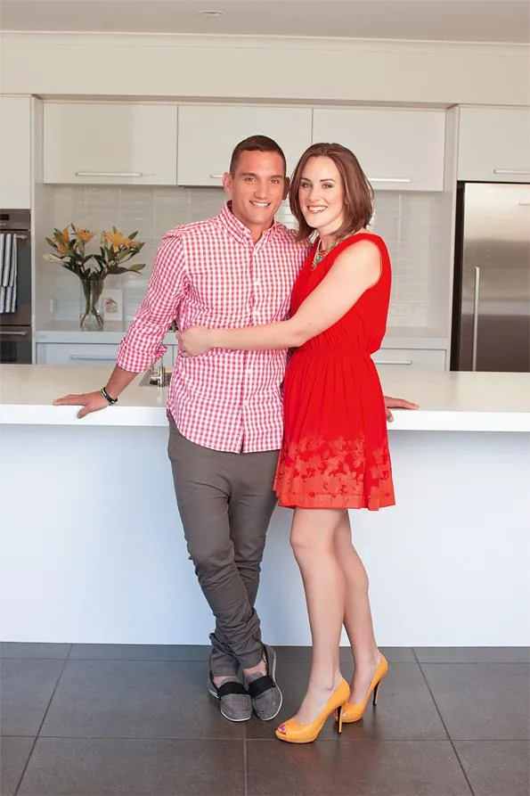 A couple stands in a modern kitchen, smiling, with the woman wearing a red dress and the man in a red checkered shirt.