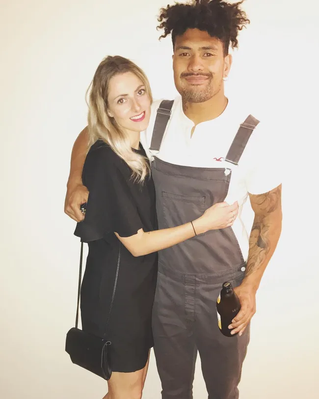 A woman in a black dress and a man in overalls with a drink pose smiling against a plain background.