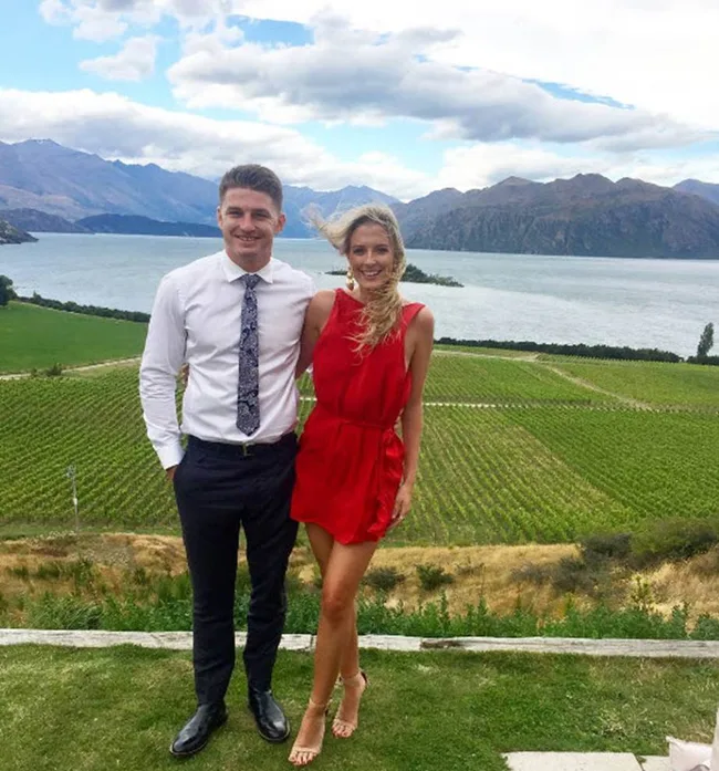A couple posing in front of a vineyard and lake, mountains in the background; the woman in a red dress, the man in a white shirt.