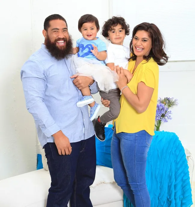 A smiling family with two children posing in a bright room, one child held by the father, both adults are joyful.