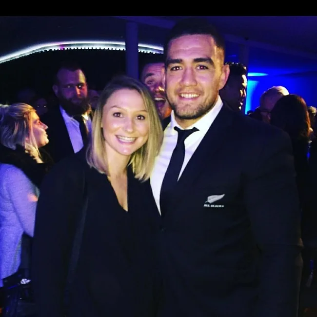 A woman and a man in a suit with a rugby logo on his blazer, smiling at a semi-formal indoor event.