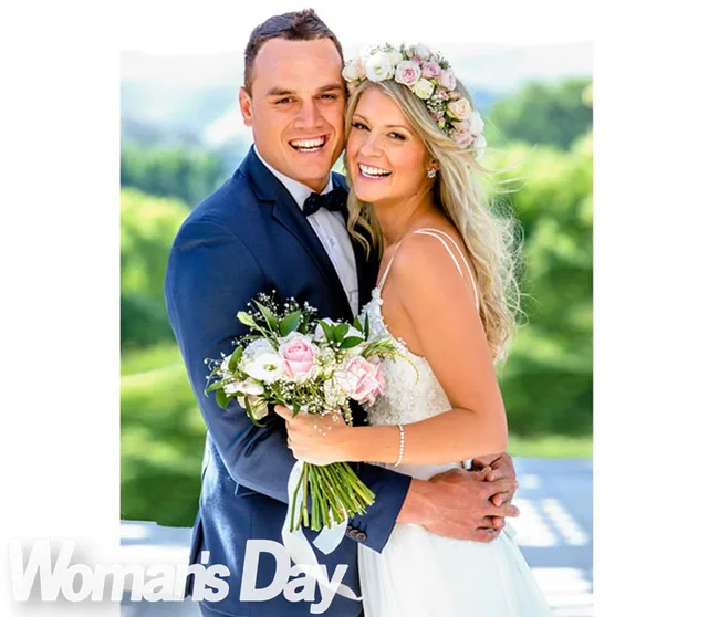 A bride in a white dress and floral crown, and groom in a blue suit, smile while holding a bouquet outdoors.
