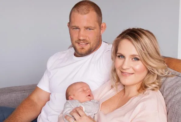 A couple sitting on a couch, smiling with a newborn baby asleep in the woman's arms.
