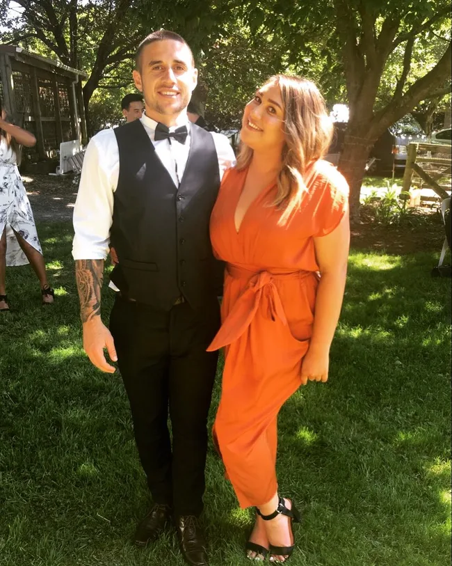 A man in formal wear and a woman in an orange dress smile while standing on grass at an outdoor event.