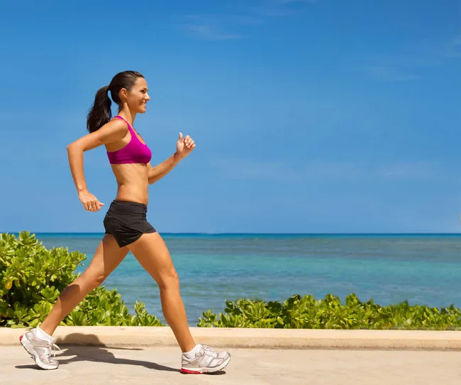 Woman jogging along a beachside path under a clear blue sky, wearing sportswear.
