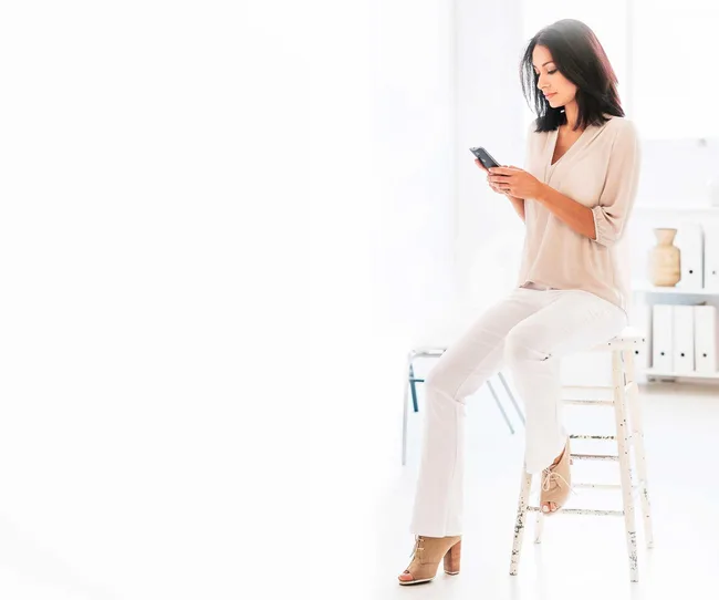 A woman sitting on a stool in a bright room, looking at her smartphone, wearing a light blouse and white pants.