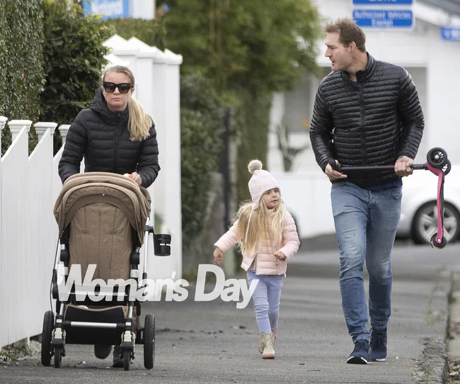A family strolls on a sidewalk, woman pushing a stroller, and man holding a scooter, with a young girl walking alongside.