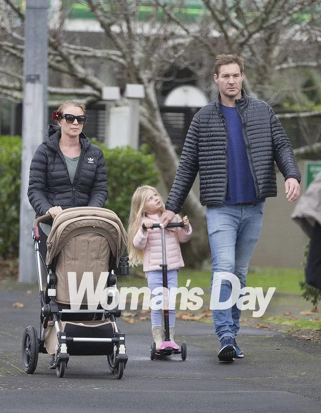 A family walks outdoors; the woman pushes a stroller, the man and child with scooter walk beside her.