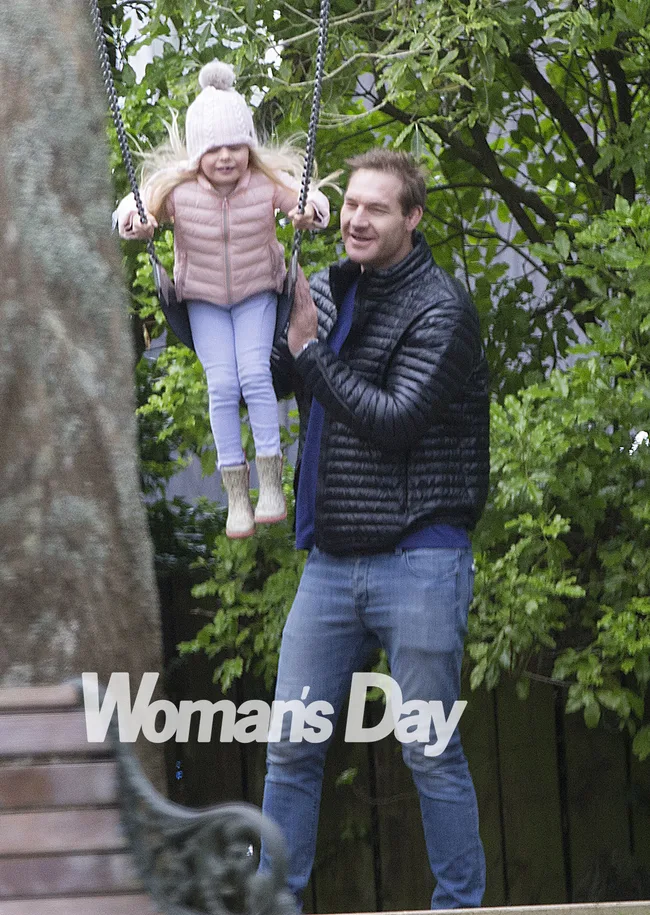 A man pushes a little girl on a swing in a park, both smiling. The girl wears a pink jacket and beanie.