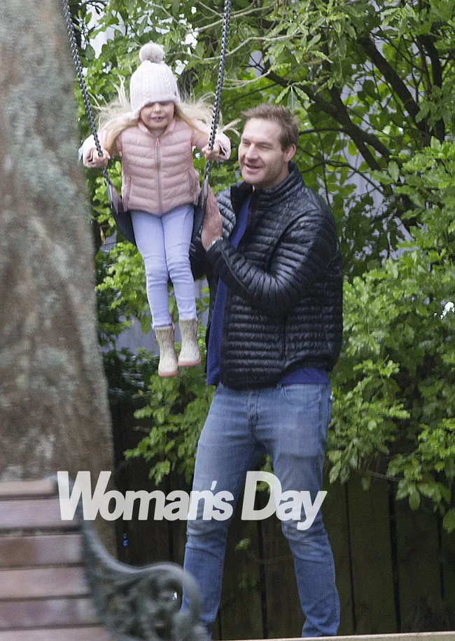 A man pushes a little girl on a swing in a park, both smiling. The girl wears a pink jacket and beanie.