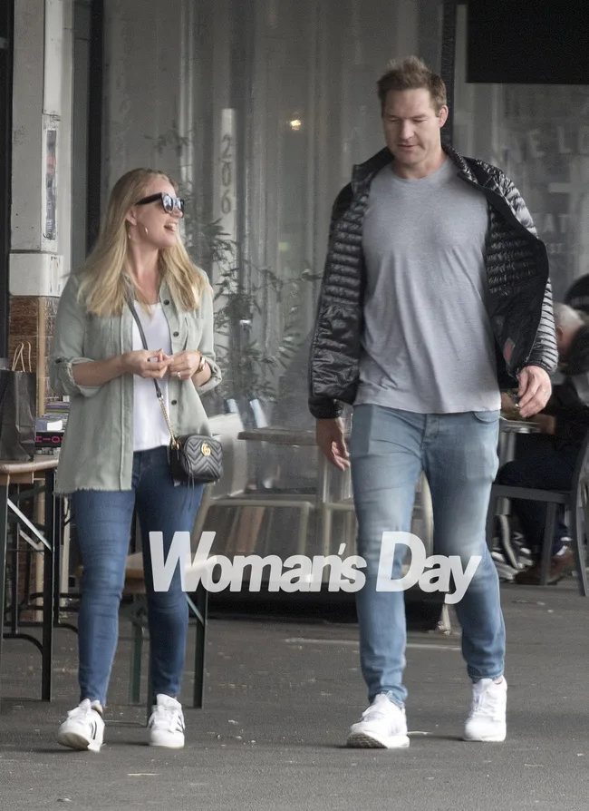 A woman and man walking on a sidewalk, both smiling, wearing casual attire with "Woman's Day" text in front.