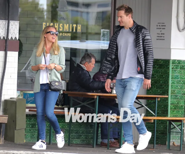 Two people walking outside a cafe, one wearing a light jacket and sunglasses, the other in a black jacket and jeans.