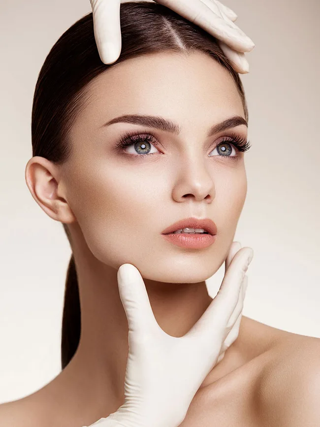 A woman with smooth skin is gently touched by gloved hands, suggesting cosmetic examination or beauty treatment.
