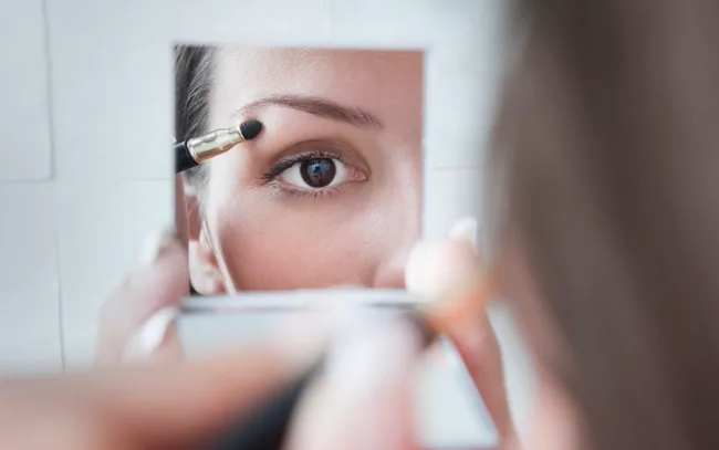 Applying makeup to eyelid using a brush, reflected in a mirror.