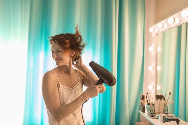 A woman in a camisole smiles while drying her hair in front of a vanity with a mirror and makeup brushes.