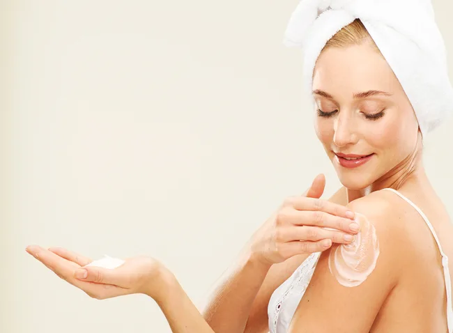 A woman applies lotion to her shoulder, wearing a towel on her head and a white tank top, smiling gently.
