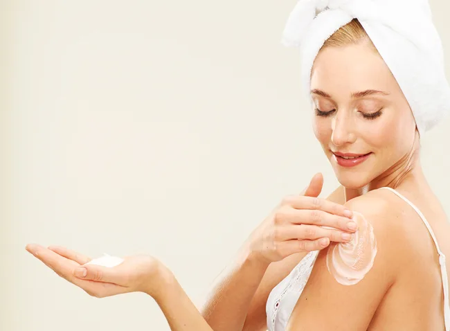 A woman applies lotion to her shoulder, wearing a towel on her head and a white tank top, smiling gently.