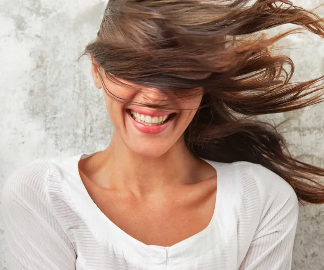 Smiling woman with windblown hair covering her eyes, wearing a white shirt against a textured background.