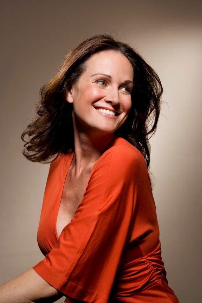Smiling woman with brunette hair in an orange top, looking to the side against a neutral background.