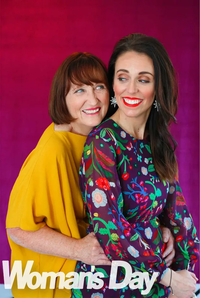 Two women smiling and embracing against a magenta background, one wearing a yellow top and the other a floral dress.