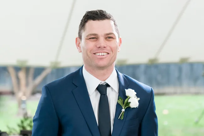A man in a blue suit with a white flower boutonniere, smiling at a wedding ceremony.
