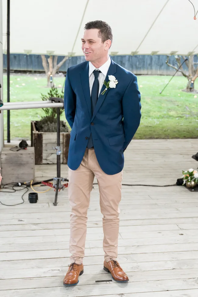 Man in blue suit with a white boutonni&egrave;re, standing on wooden floor outdoors.