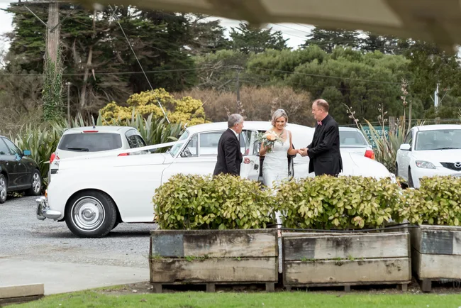 Wedding scene with a bride holding flowers exiting a white vintage car, assisted by two men in suits.
