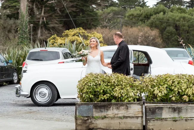 Bride in a white dress holding a bouquet, standing by a white classic car with a man in a suit, outdoors.