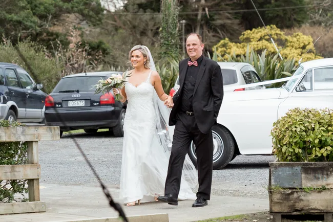 Bride in white gown holding bouquet, walking with man in black suit, outdoors near parked cars and greenery.