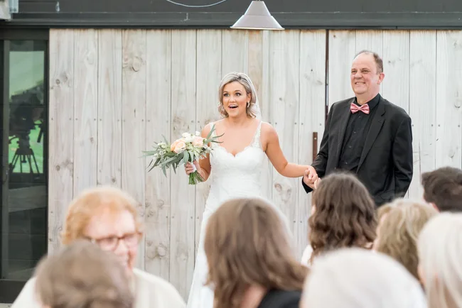 Bride in white gown holding flowers and smiling, walking with a man in black suit and pink bow tie, in front of seated guests.