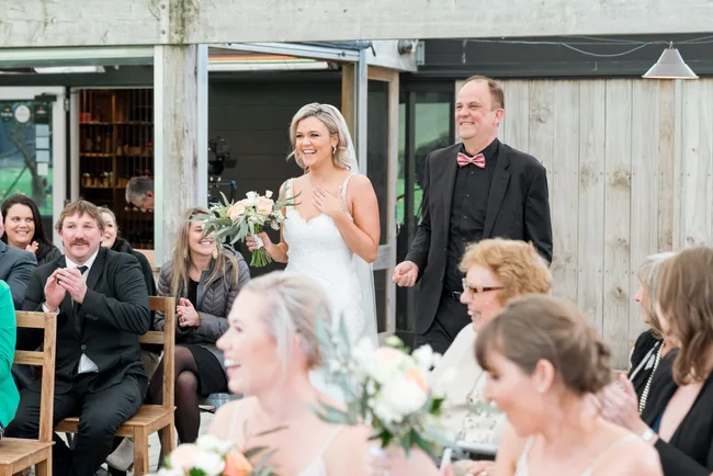 A bride in a white dress smiles while walking with a man in a pink bow tie as guests clap at a wedding reception.