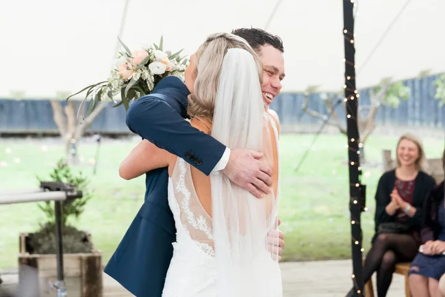 Bride and groom embrace joyfully under a tent; guests clapping in the background.