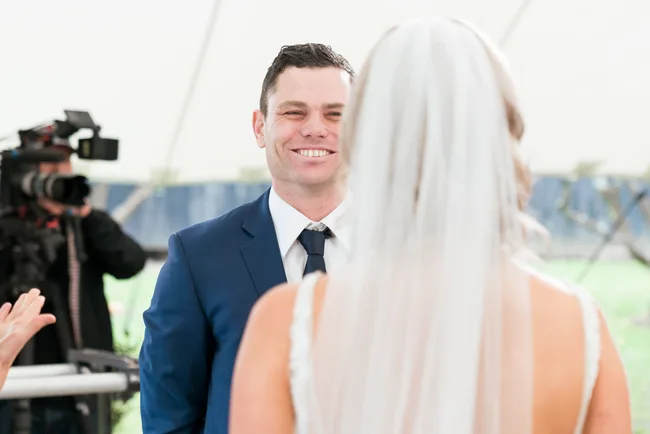 "Smiling groom in blue suit faces bride with long veil, while cameraman films the wedding ceremony."