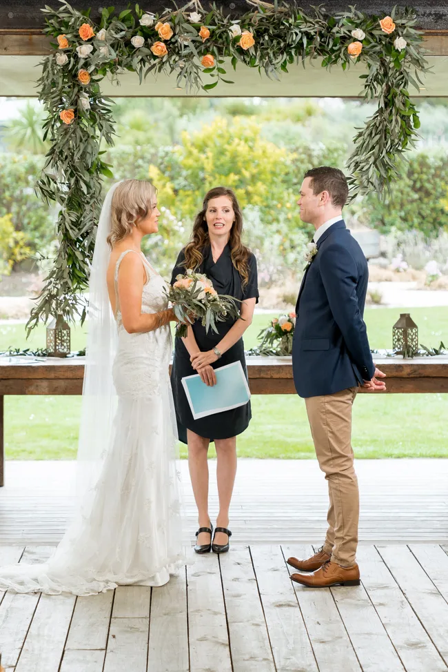 Bride and groom stand before an officiant under a floral arch during their wedding ceremony.