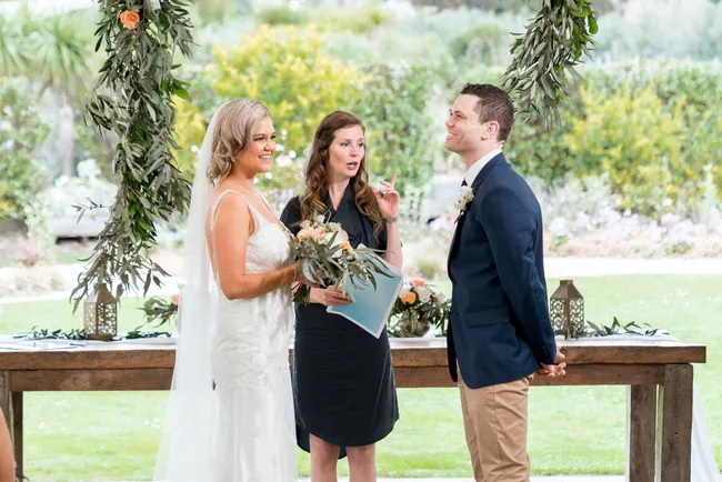 A bride and groom smile at each other during an outdoor wedding ceremony with an officiant between them.