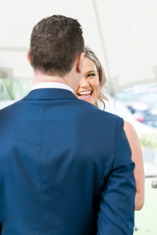 Two people at a wedding ceremony. A woman smiling at a man in a blue suit, with cars in the background.
