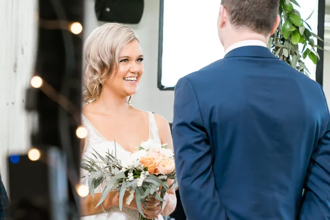 Bride smiling at groom, holding bouquet, during wedding ceremony indoors.