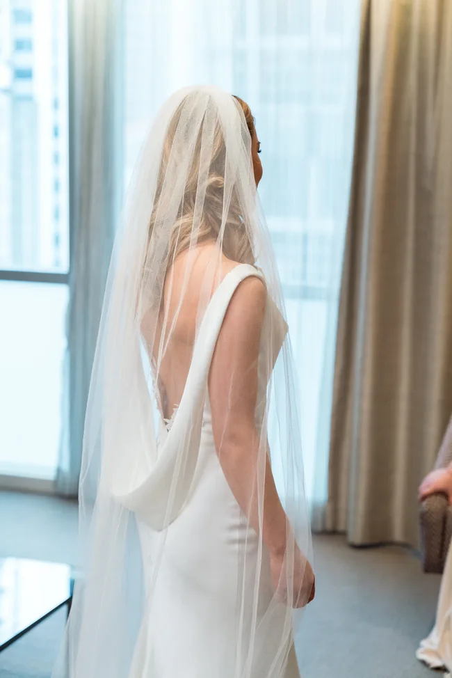 Bride in a white wedding dress and veil, standing indoors near a window with sheer curtains.