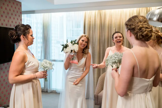 Bridesmaids in light dresses surround a smiling bride holding a bouquet in a bright room.