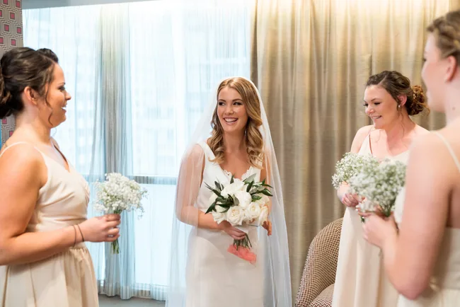 Bride in white dress holding bouquet with bridesmaids in light dresses, smiling in a room with curtains.