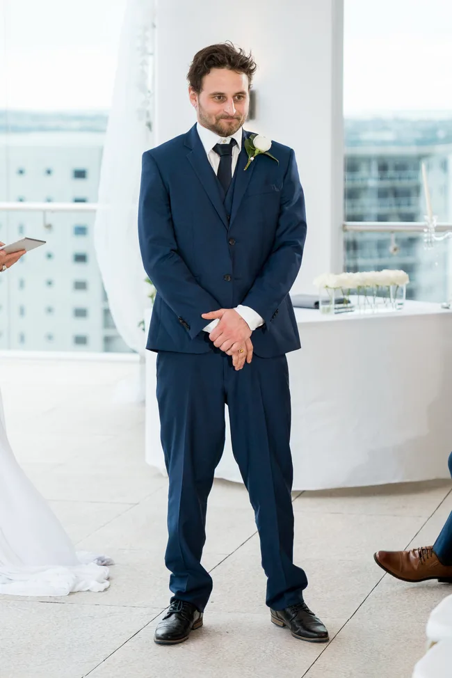 Man in a blue suit with a boutonniere standing, looking forward. White backdrop with large windows and city view.
