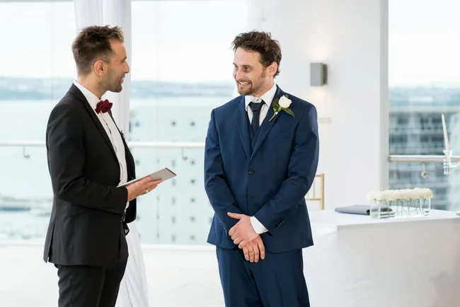 Two men in suits at a wedding ceremony, one holding a tablet, the other smiling with a boutonniere.
