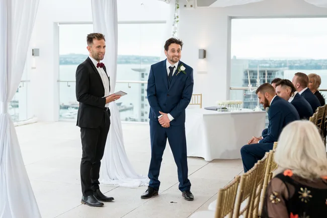 Man in suit stands at altar during wedding ceremony, smiling, with officiant and guests seated nearby.