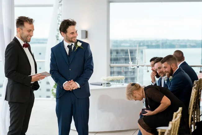 Best man and groom in blue suits smiling at a wedding ceremony indoors.